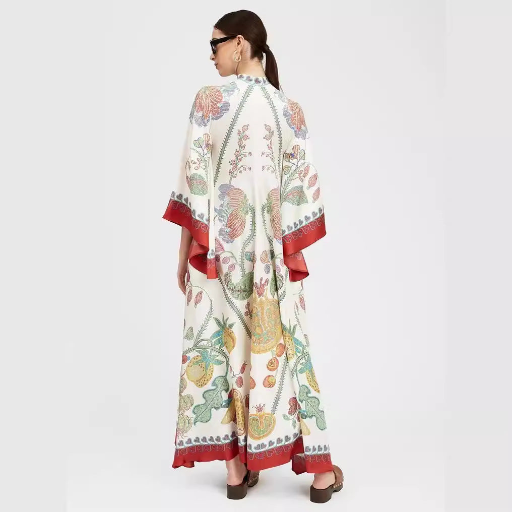 Back view of an off-white floor-length maxi dress featuring a vibrant botanical fruit and floral print, flared 3/4 sleeves, and red decorative border trim along the sleeve cuffs and hem.