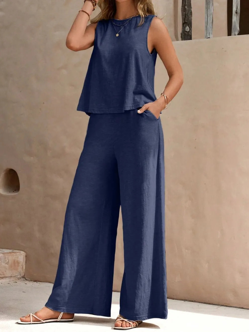 Front view of model with hands in pockets wearing a dusty blue sleeveless round neck top and high-waisted wide leg pants set with sandals