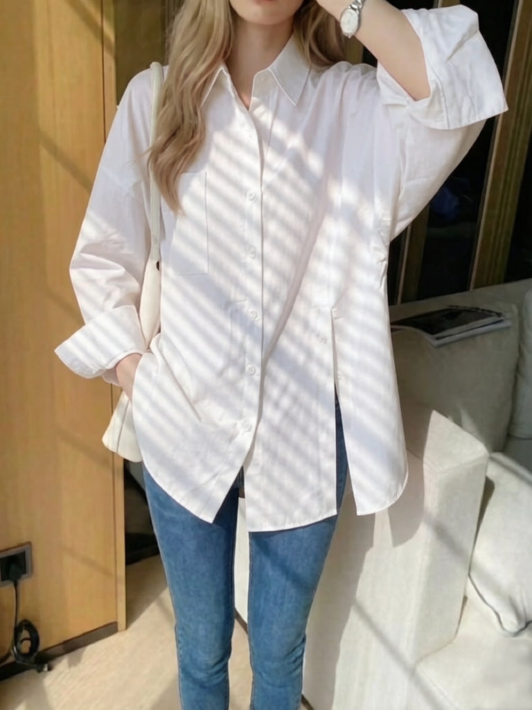 Woman wearing a white oversized long-sleeve button-down shirt with high side slits, styled with blue jeans in bright window sunlight.