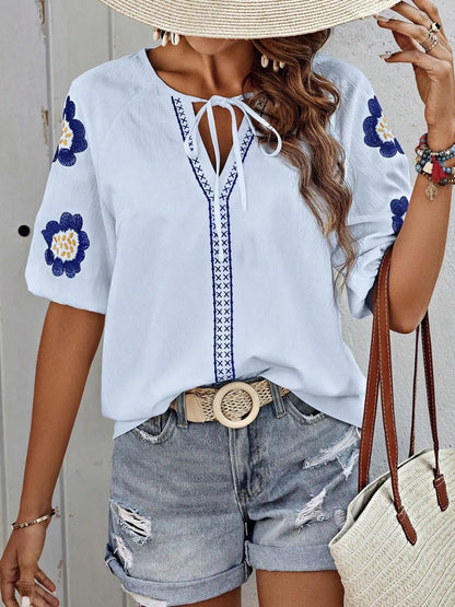 Woman wearing a light blue V-neck blouse with embroidered flower sleeves, a braided tie detail, and distressed denim shorts.