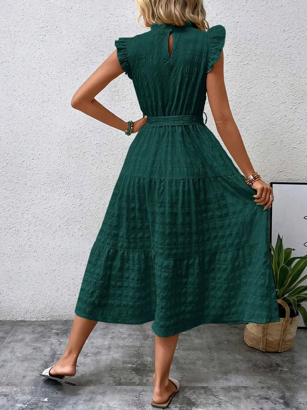 Back view of model wearing a dark green ruffled cap sleeve midi dress with a tied waist and white slide sandals