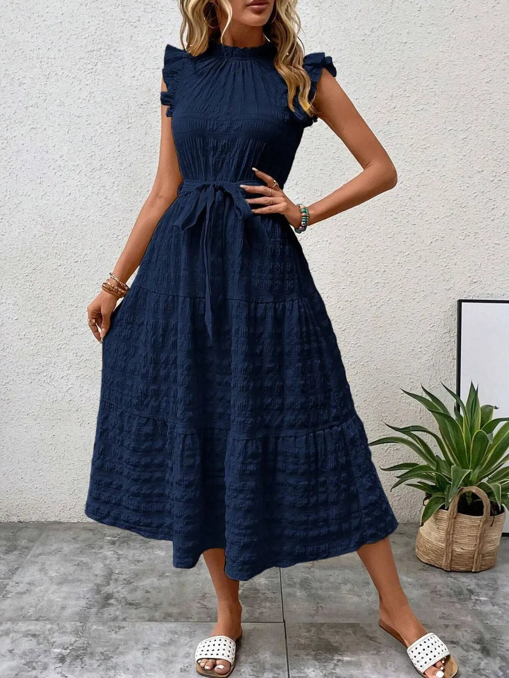 Front view of a model wearing a dark navy blue ruffled cap sleeve midi dress with a tied waist and white slide sandals.
