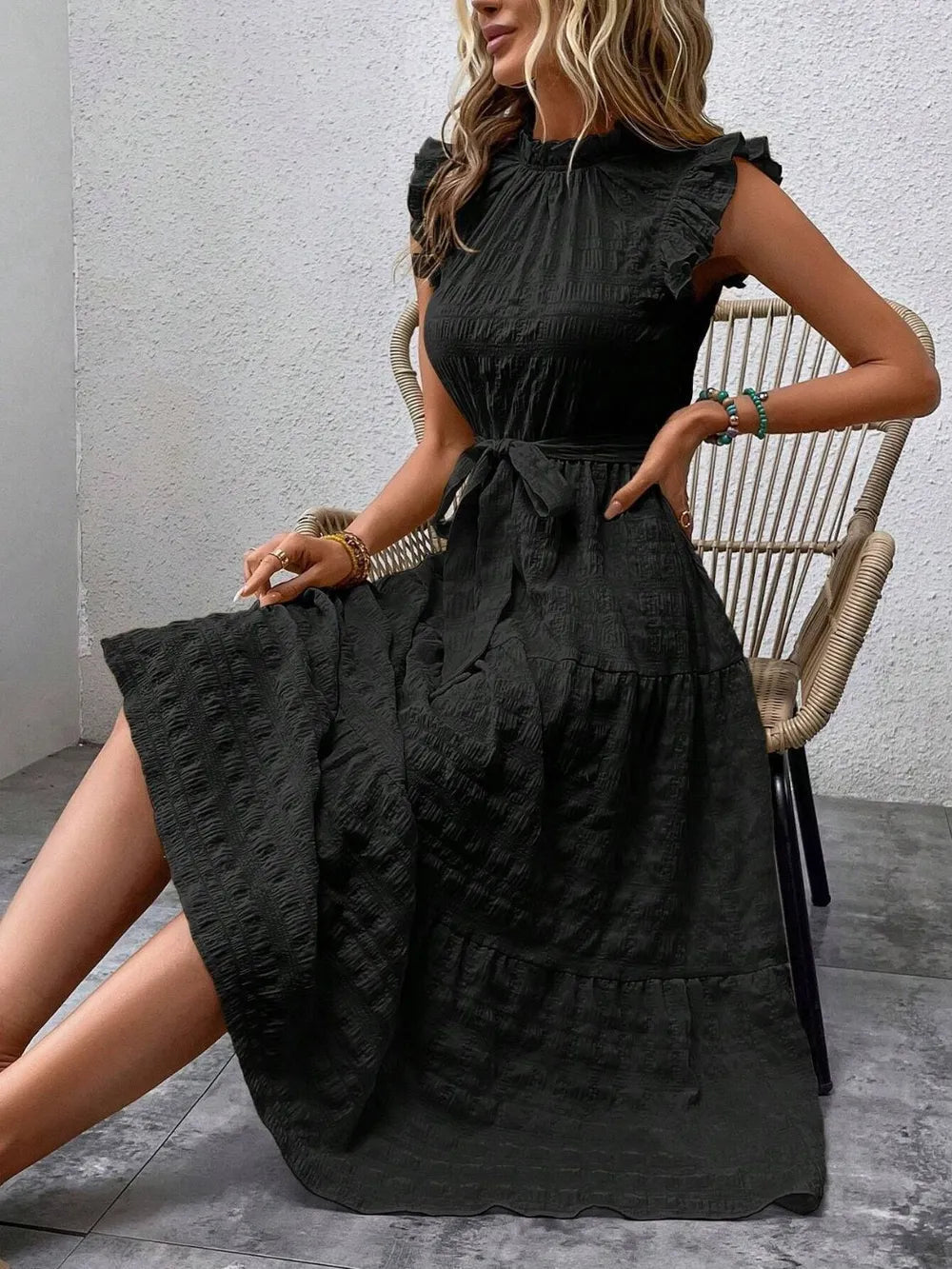 Model sitting on a woven chair wearing a black ruffled cap sleeve midi dress with a tied waist.