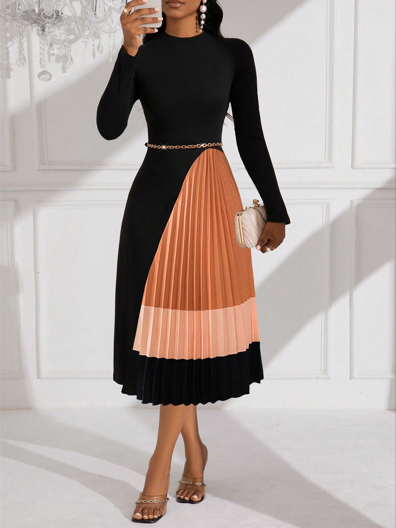 Model wearing the Lyxana long-sleeve mock neck midi dress featuring a fitted black top, gold waist chain, and a colorblock beige and burnt orange pleated skirt, paired with gold heels and holding a small white clutch