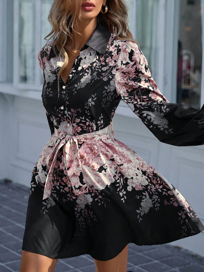 A medium close-up view of a woman wearing a black mini shirt dress with a pink floral print, featuring a grey collar, front button closure, volumetric bishop sleeves, and a matching patterned belt tied around the waist.