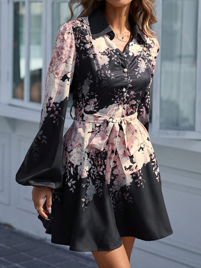 A mid-section view of a woman wearing a black mini shirt dress with a pink floral print, featuring a lapel collar, button-up front, wide bishop sleeves, and a matching fabric waist belt tied at the front.