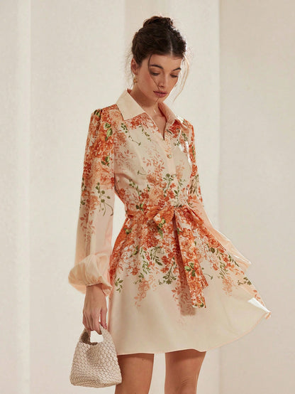A full-length photo of a woman wearing a cream-colored shirt dress with an orange and green floral print, featuring long bishop sleeves, a lapel collar, a tied waist belt, and carrying a small textured woven handbag.
