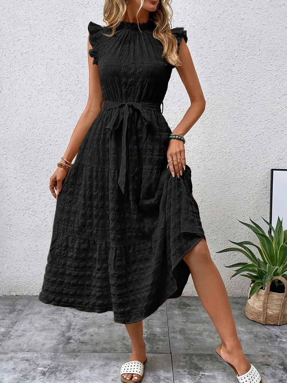 Front view of a model wearing a black ruffled cap sleeve midi dress with a tied waist and white slide sandals.