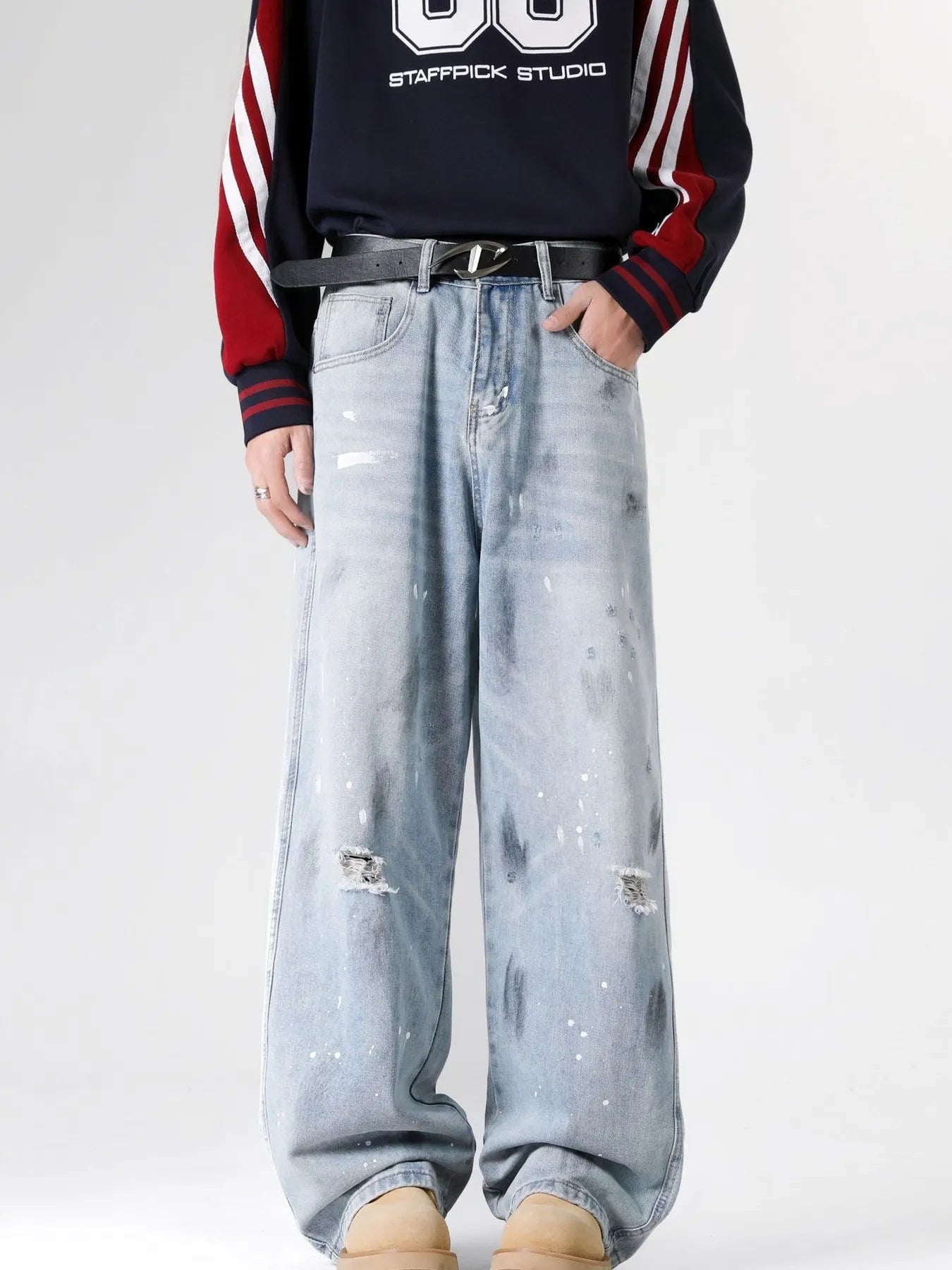 Front view of a person wearing light blue distressed wide-leg jeans with paint splatter details, paired with a dark sweatshirt featuring red and white sleeve stripes, against a dark background.
