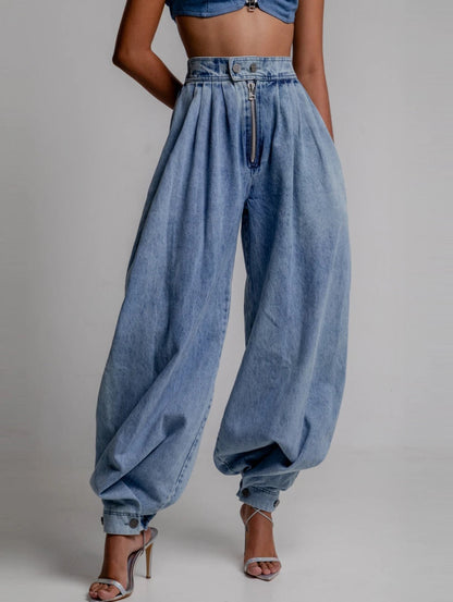 Close-up detail view of high-waisted light blue denim harem pants showing the exposed front zipper, button waist closure, pleated front construction, and relaxed fit against a neutral background.