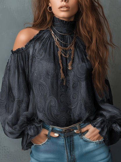 Woman wearing a dark gray retro cashew print off-shoulder blouse with a high ruffled mock neck, dramatic lantern sleeves, and layered pendant necklaces, styled with blue denim jeans.