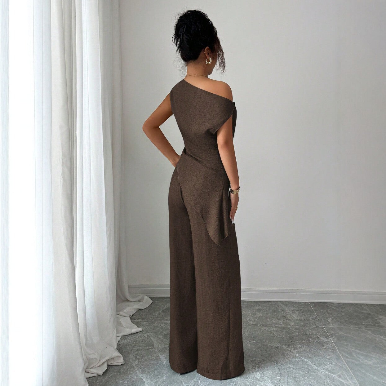 Woman seen from the side wearing a chocolate brown casual two piece set featuring an asymmetric one-shoulder top and matching high-waisted wide-leg pants.