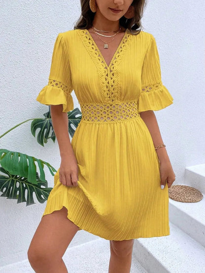 Woman wearing a bright yellow V-neck mini dress with textured vertical striping, featuring crochet lace trim along the V-neckline, waist cutouts, and flounce sleeve cuffs, styled with layered gold pendant necklaces.