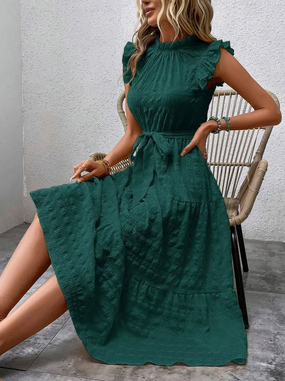 Model sitting on a woven chair wearing a dark green ruffled cap sleeve midi dress with a tied waist