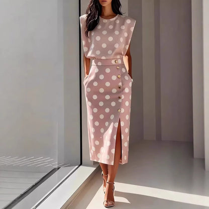 Model standing near a window wearing a pink polka-dot sleeveless top and matching high-slit maxi skirt set with strappy heels.