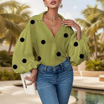 Woman wearing a loose matcha green top with black polka dots, featuring an open V-neck collar, voluminous long bubble sleeves, gold hoop earrings, layered necklaces, tucked into high-waisted blue jeans while holding a white clutch outdoors near a pool.