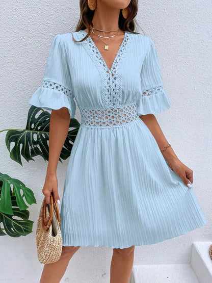 Woman wearing a light blue V-neck mini dress with textured vertical striping, featuring crochet lace trim along the V-neckline, waist cutouts, and flounce sleeves, holding a woven straw tote bag.