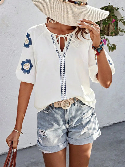 Woman wearing a white V-neck blouse with blue embroidered flowers on the sleeves, braided tie neck detail, and intricate blue-and-white patterned trim along the neckline, styled with distressed cuffed denim shorts, a woven belt, and a wide-brim straw hat.