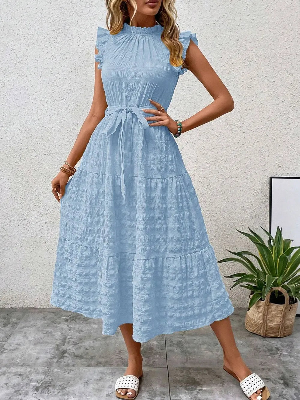 Front view of model wearing a light blue ruffled cap sleeve midi dress with a tied waist and white slide sandals