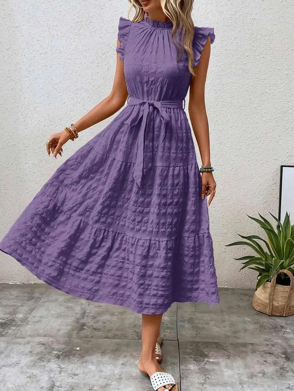 Front view of a model stepping forward wearing a purple ruffled cap sleeve midi dress with a tied waist and white slide sandals.