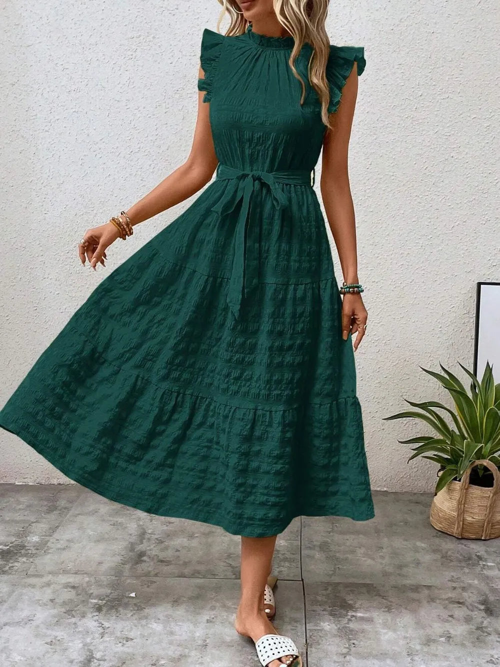 Front view of model stepping forward wearing a dark green ruffled cap sleeve midi dress with a tied waist and white slide sandals