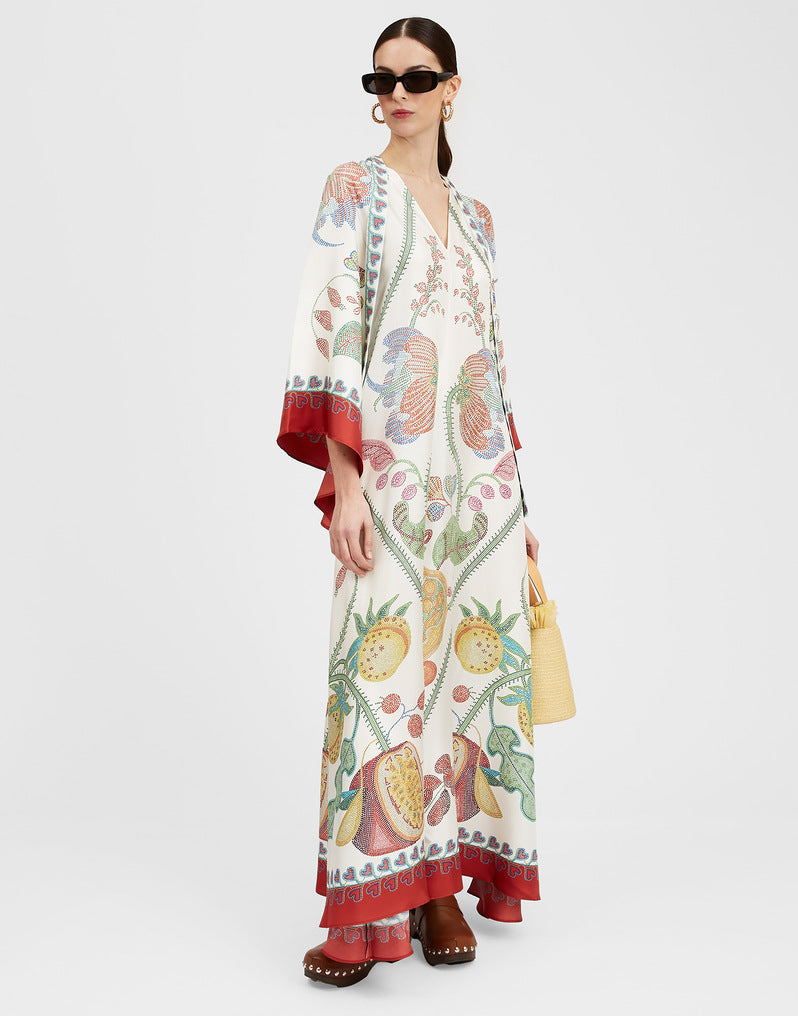 Women's printed floor-length A-line maxi dress in off-white featuring a tropical fruit and botanical pattern, V-neckline, wide 3/4 flared sleeves, and red decorative border trim.