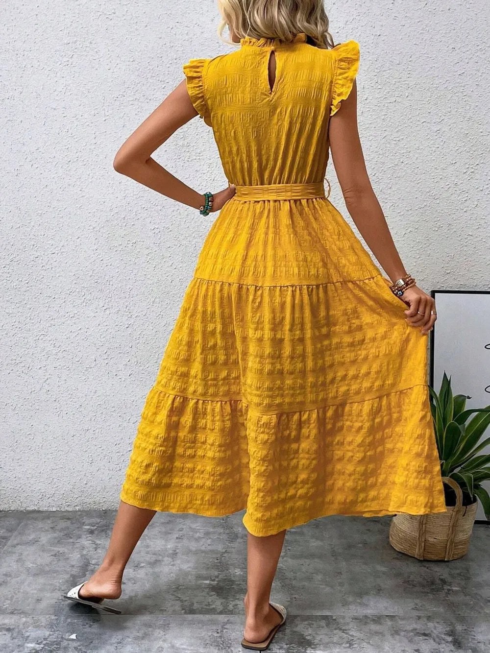 Back view of model wearing a gold yellow ruffled cap sleeve midi dress with a tied waist and white slide sandals