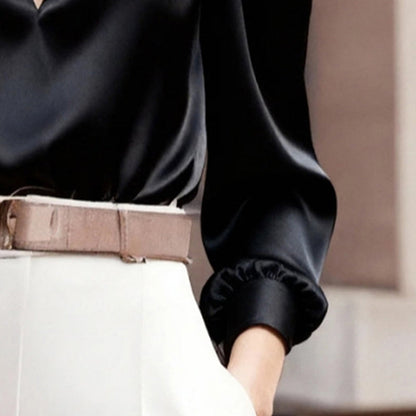 Close-up of a woman wearing a black satin V-neck long-sleeve blouse tucked into white trousers accented with a beige belt.