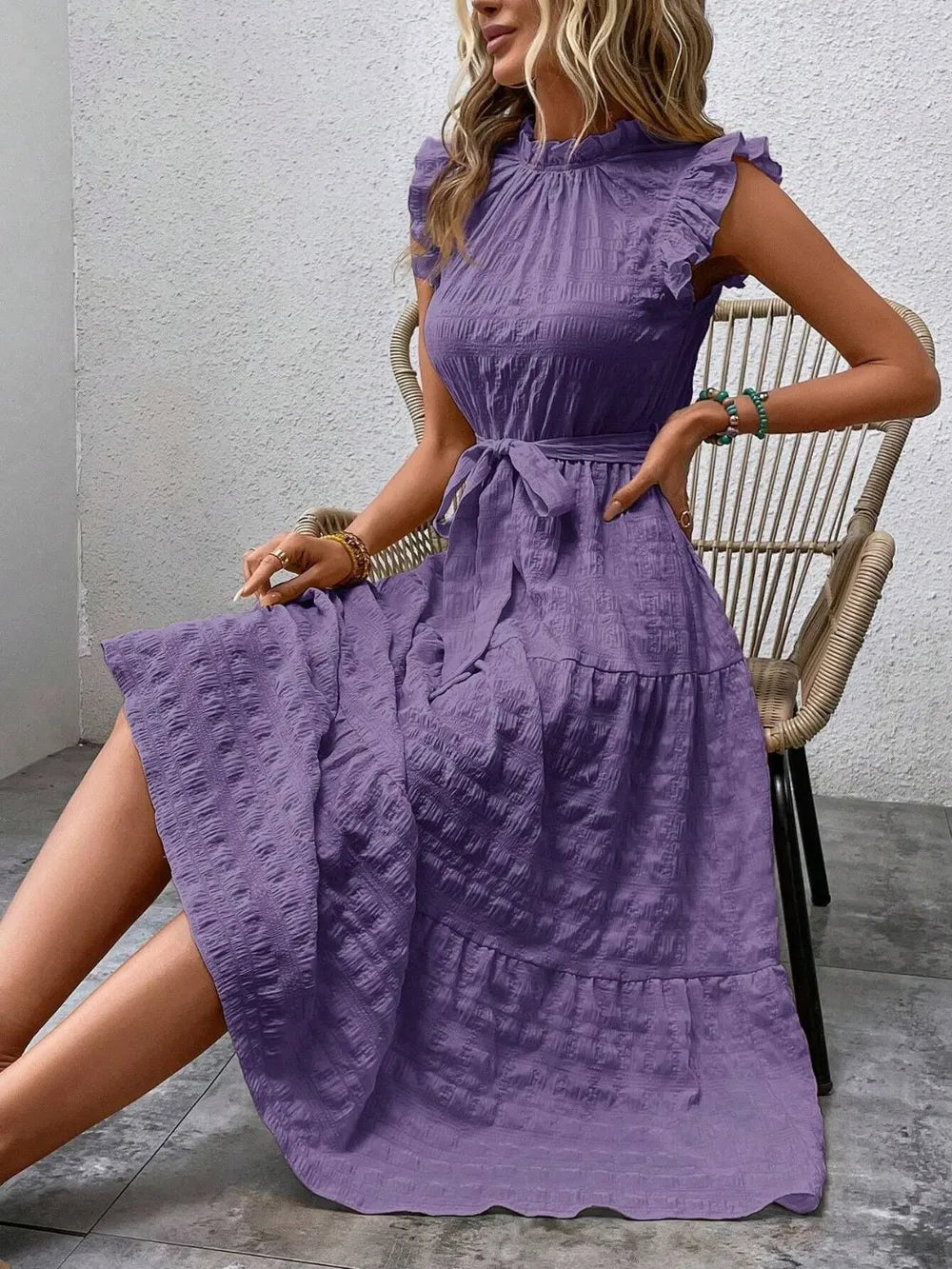 Model sitting on a woven chair wearing a purple ruffled cap sleeve midi dress with a tied waist.
