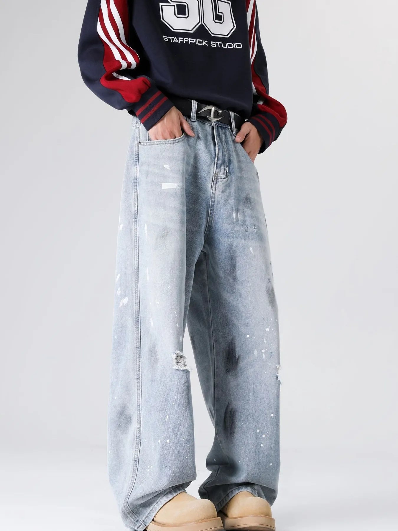Lower-body view of a person wearing light blue distressed wide-leg jeans with paint splatter details and tan footwear, against a dark background.