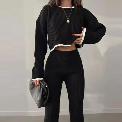 Woman wearing a black ribbed knit two-piece set featuring a long-sleeve crop top and matching slim pants with white trim detail, holding a grey clutch.