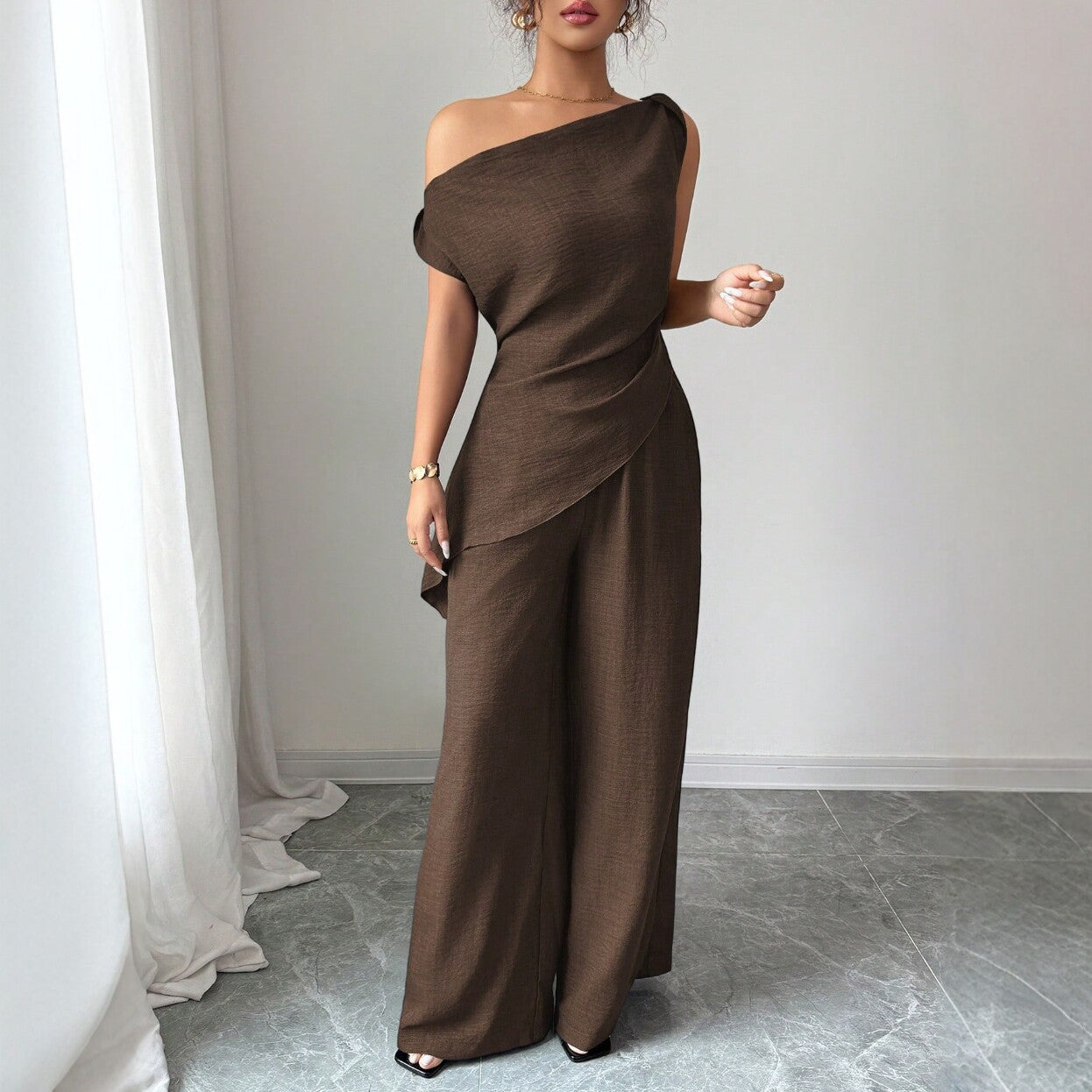 Woman wearing a dark brown casual two-piece set featuring a draped off-the-shoulder top with a layered cross-body fold and matching high-waisted wide-leg pants.