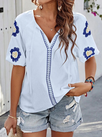 Woman wearing a light blue V-neck top with blue and yellow floral embroidered sleeves and trim, paired with distressed denim shorts.