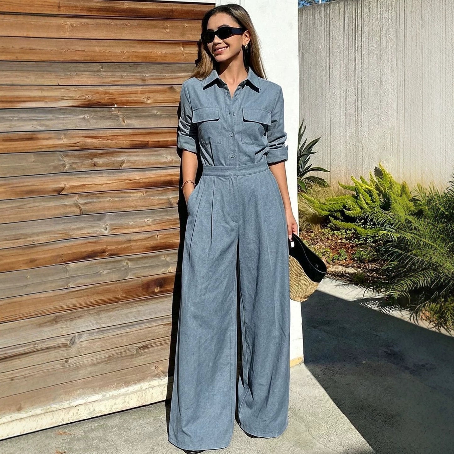 Full-length outdoor shot of a woman wearing a blue-gray collared long-sleeve jumpsuit with chest flap pockets, sunglasses, and wide-leg trousers while holding a straw clutch.