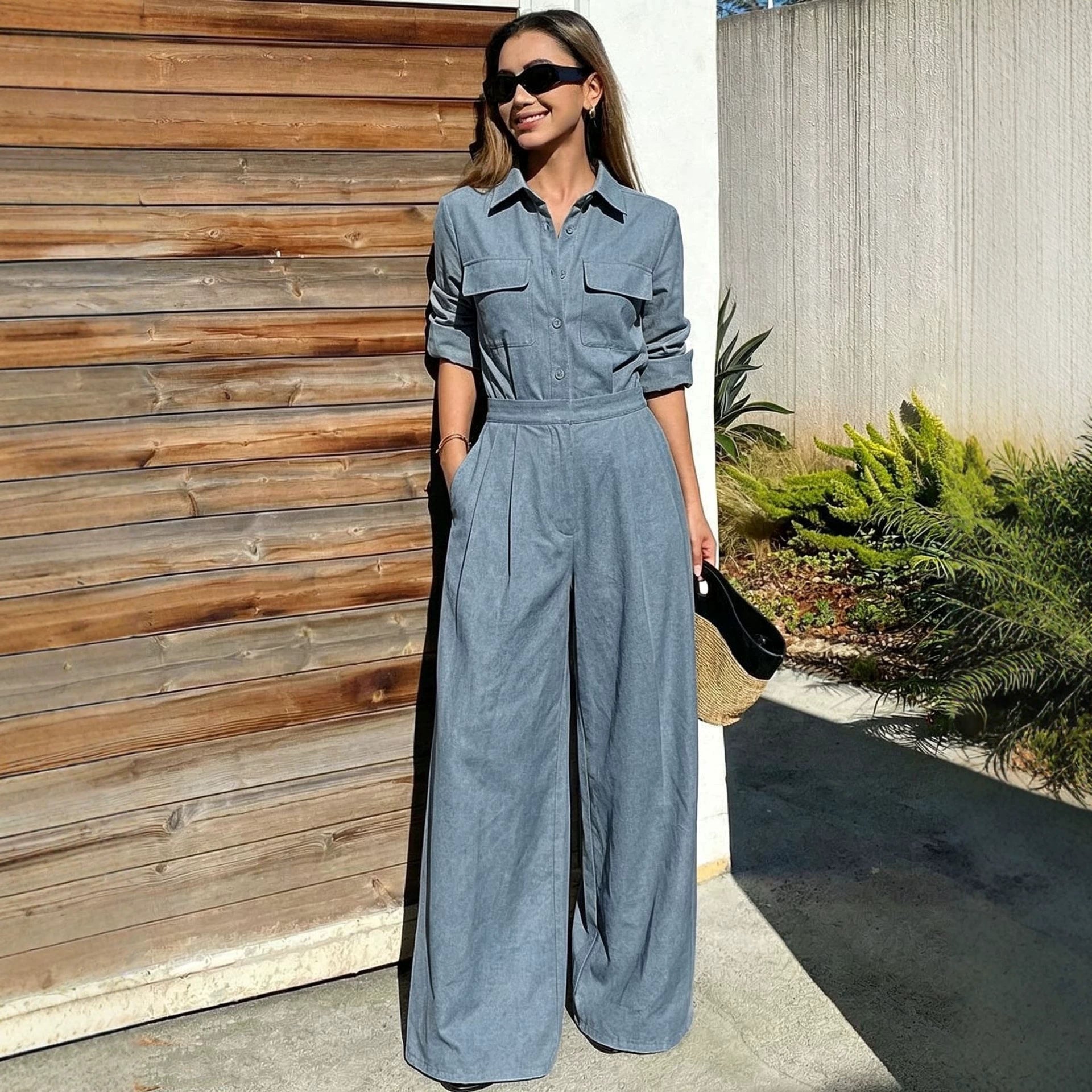 Full-length outdoor shot of a woman wearing a blue-gray collared long-sleeve jumpsuit with chest flap pockets, sunglasses, and wide-leg trousers while holding a straw clutch.