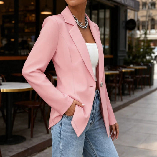 Woman wearing a pink blazer, white top, and blue jeans, standing outdoors on a city sidewalk beside a café.
