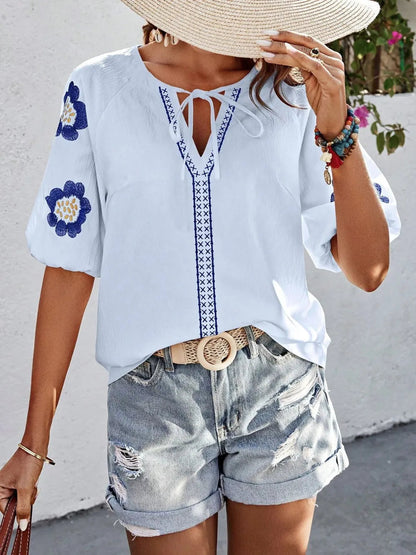 Woman wearing a straw hat and light blue V-neck top with blue floral embroidered sleeves, braided tie collar, and cuffed denim shorts.