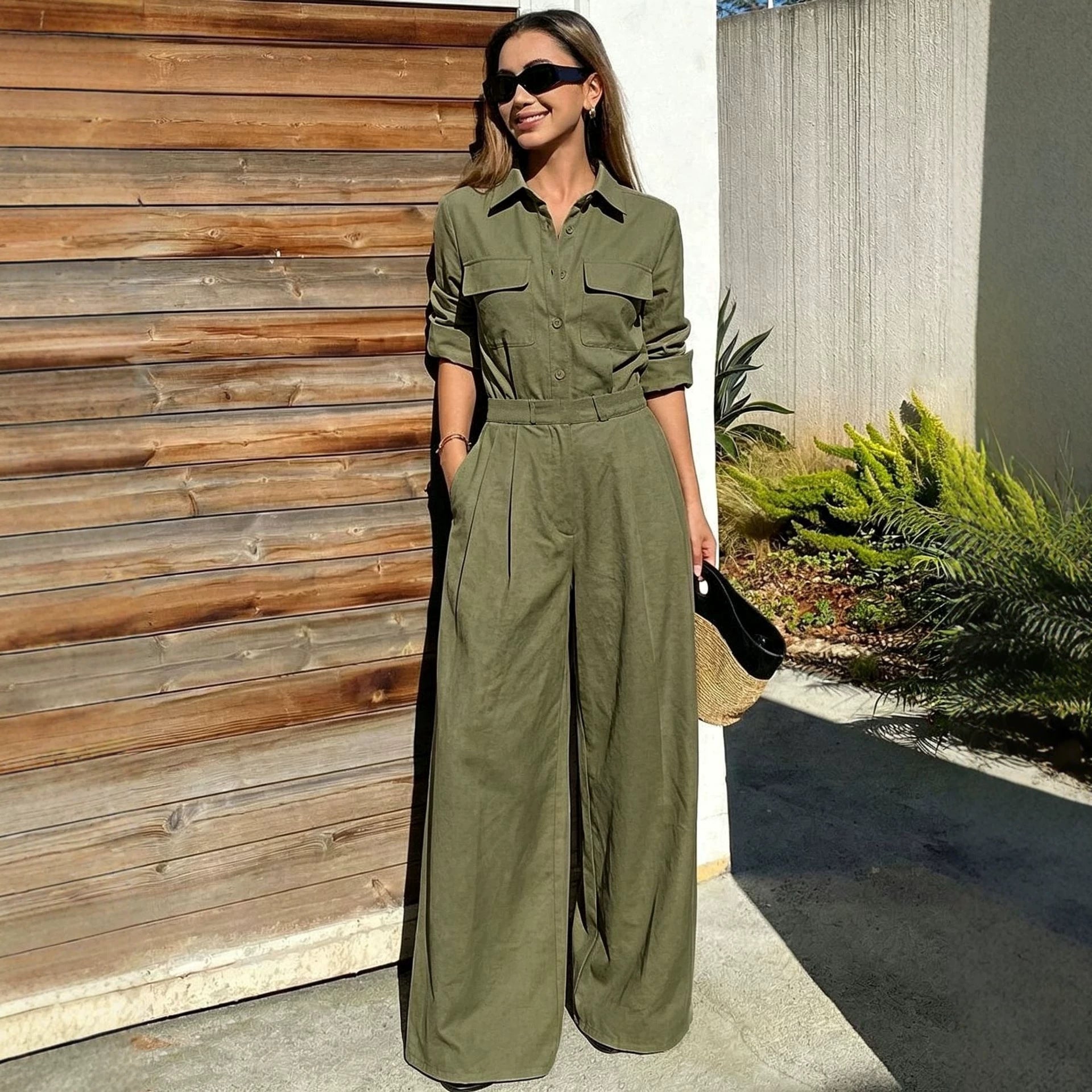 Full-length outdoor shot of a woman wearing an olive green collared long-sleeve jumpsuit with chest flap pockets, sunglasses, and wide-leg trousers.