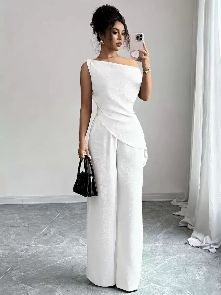 Woman wearing a white casual two-piece set featuring a draped off-the-shoulder top with a diagonal front fold and matching high-waisted wide-leg pants, holding a black handbag.