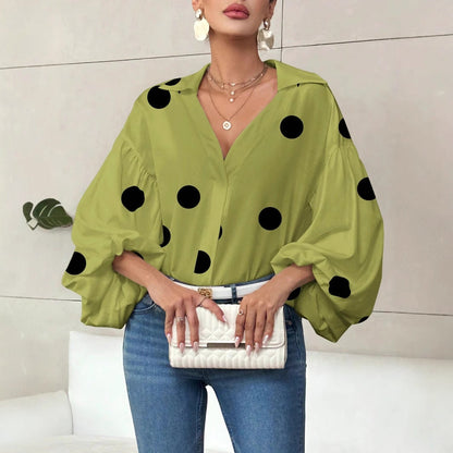 Woman wearing a loose-fit matcha green long-sleeve top with black polka dots, featuring a V-neck open collar, voluminous bubble sleeves, styled tucked into high-waisted blue jeans with a white belt, and holding a white quilted clutch.