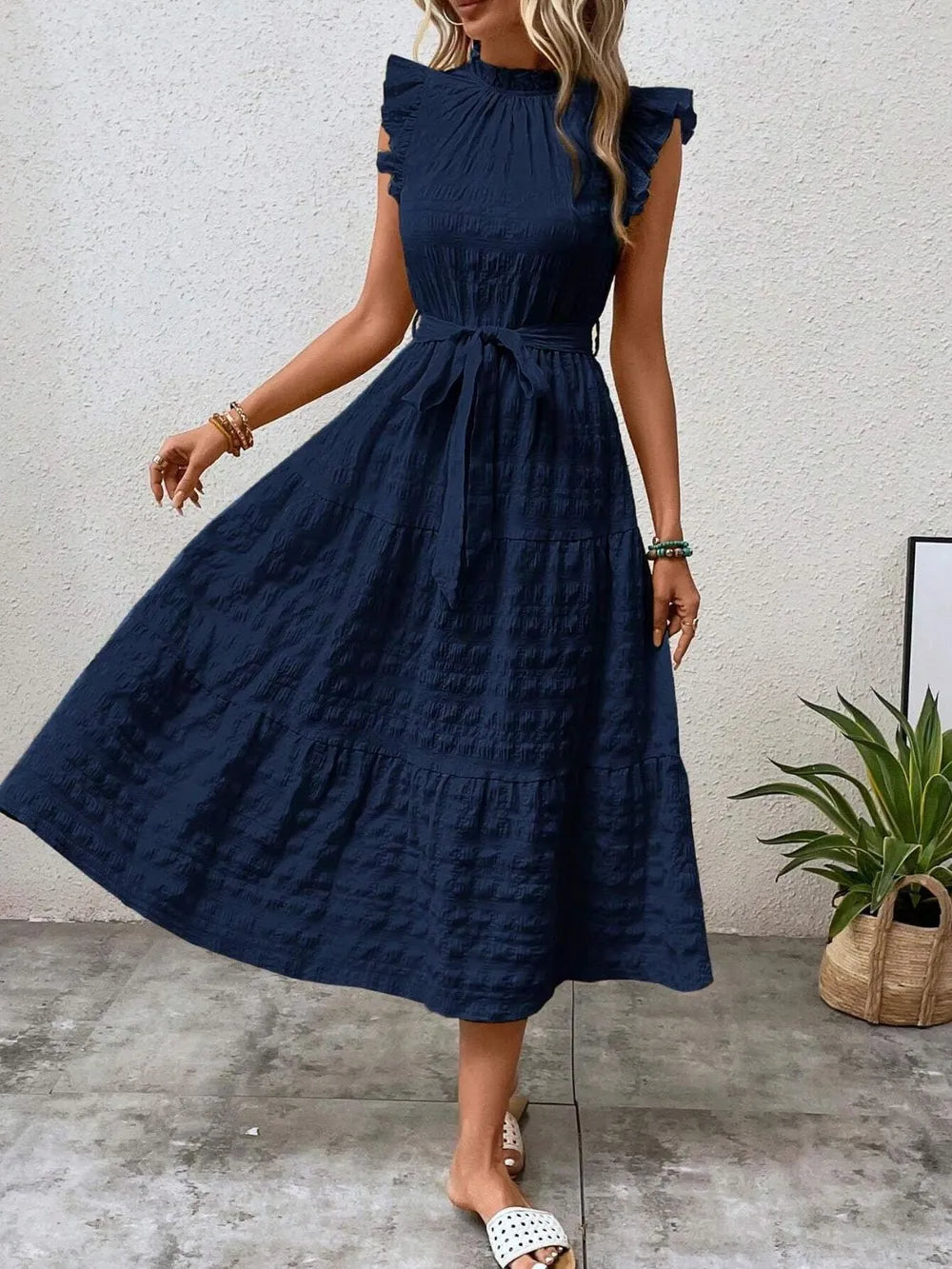 Front view of a model stepping forward wearing a dark blue ruffled cap sleeve midi dress with a tied waist and white slide sandals.