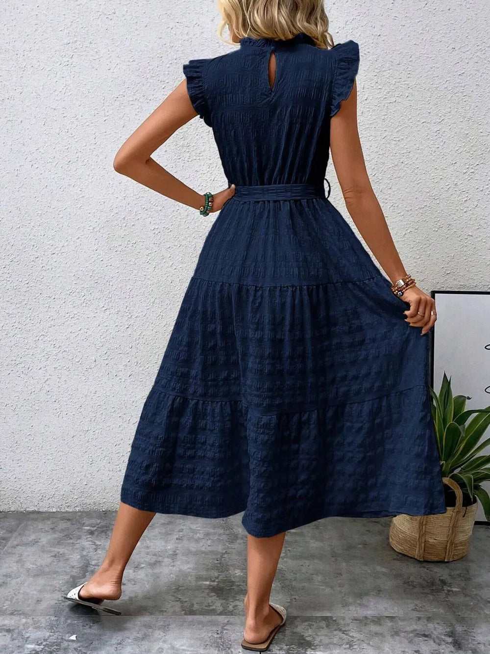Back view of a model wearing a dark blue ruffled cap sleeve midi dress with a tied waist, high neckline with keyhole detail, and white slide sandals.