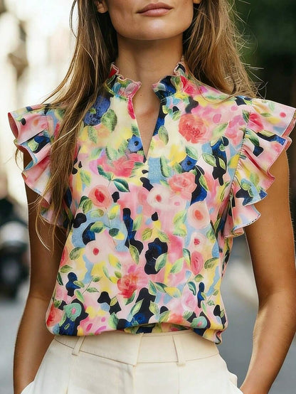 Woman wearing a multicolor floral print sleeveless top with a high ruffle-trim mock neck and lotus-leaf ruffle sleeves, styled with white high-waist pants.