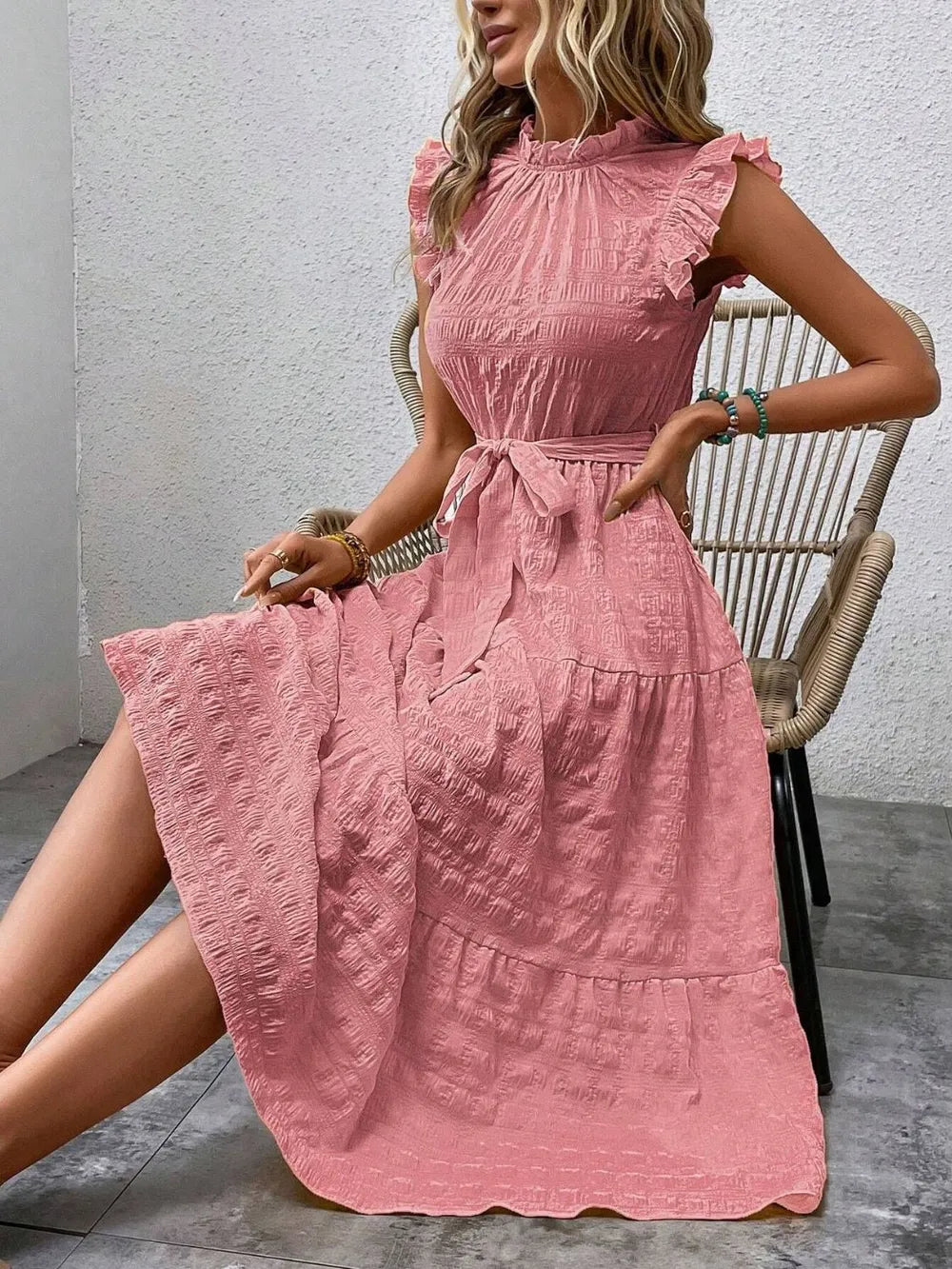 Model sitting on a woven chair wearing a pink burnt coral ruffled cap sleeve midi dress with a tied waist.
