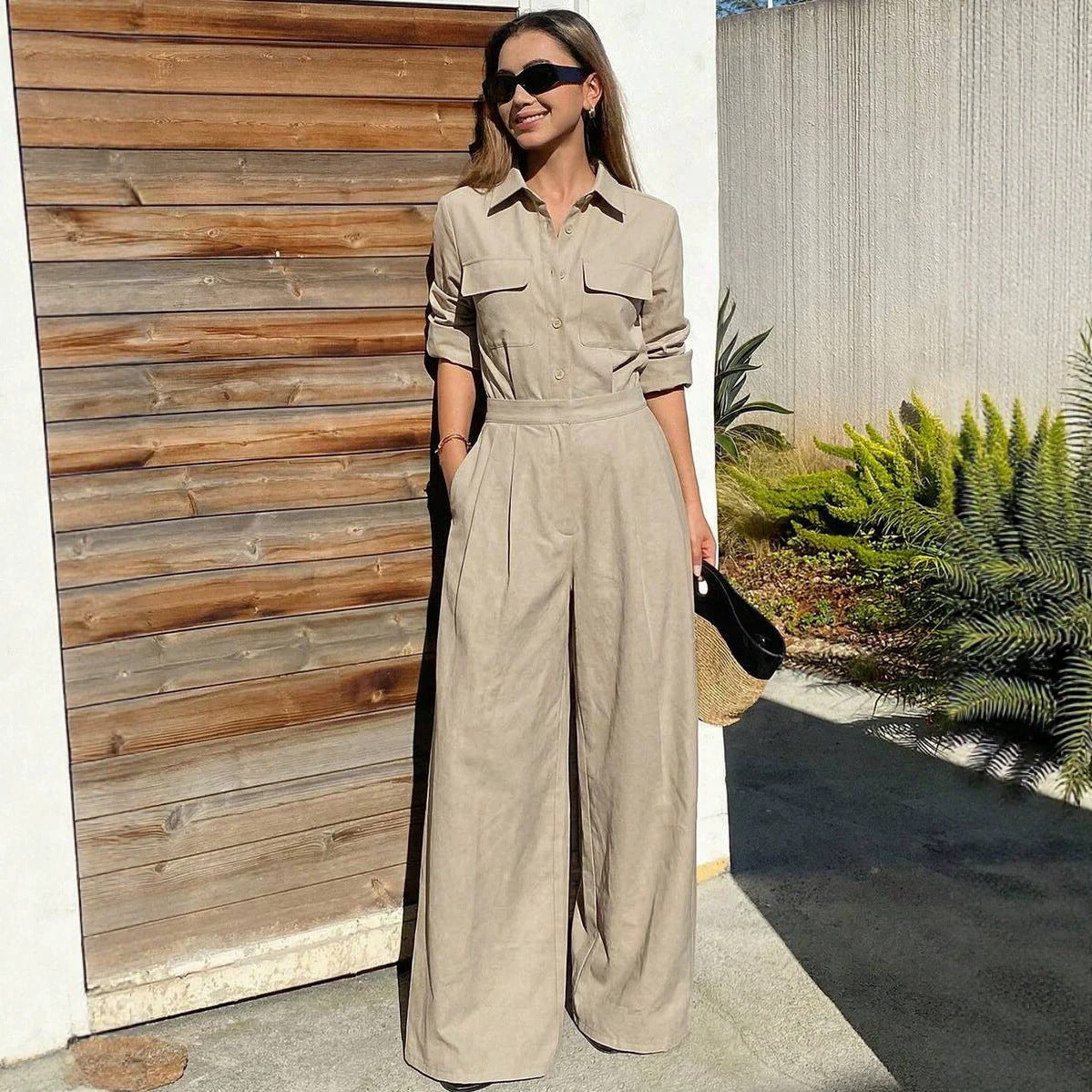 Full-length outdoor shot of a woman wearing a beige/khaki collared long-sleeve jumpsuit with chest flap pockets, sunglasses, and wide-leg trousers.