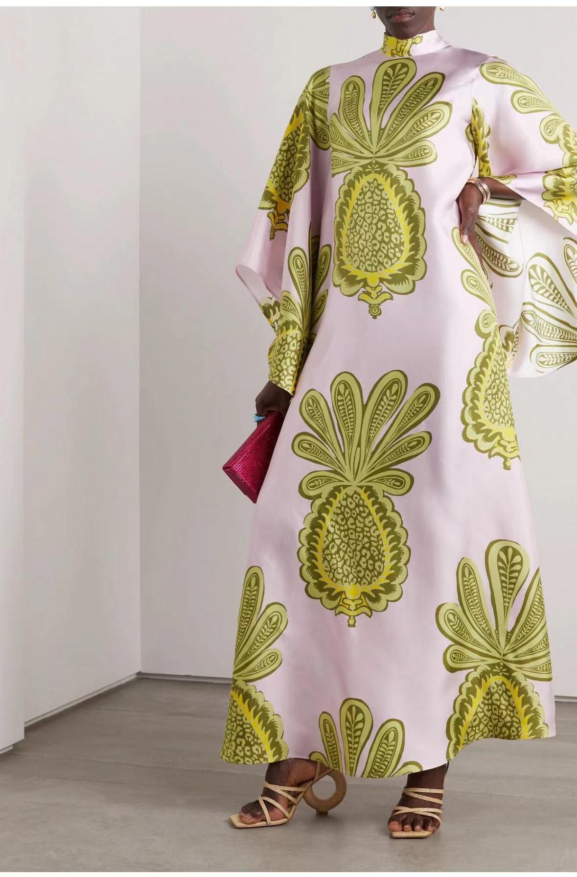 Full-length model shot of a soft pink satin maxi dress featuring a high mock stand collar, wide 3/4 flared sleeves, and a large olive-green and gold stylized botanical pineapple-motif print.
