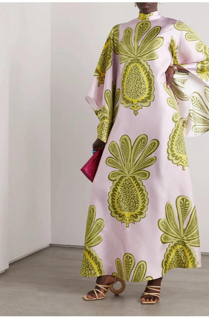 Full-length model shot of a soft pink satin maxi dress featuring a high mock stand collar, wide 3/4 flared sleeves, and a large olive-green and gold stylized botanical pineapple-motif print.