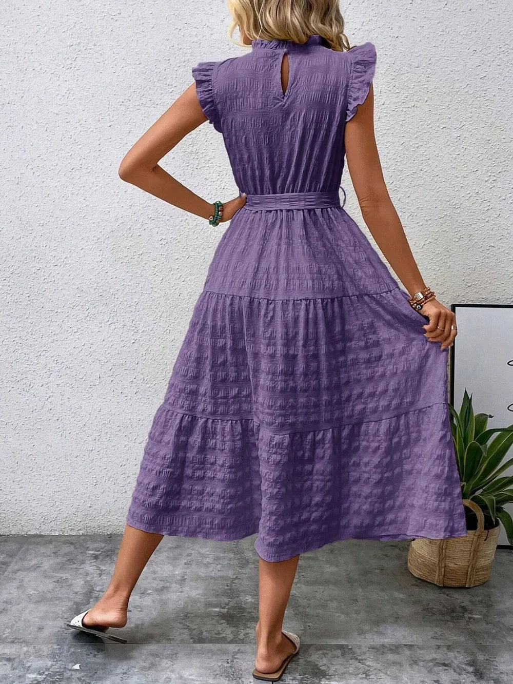 Back view of a model wearing a purple ruffled cap sleeve midi dress with a tied waist, high neckline with keyhole detail, and white slide sandals.