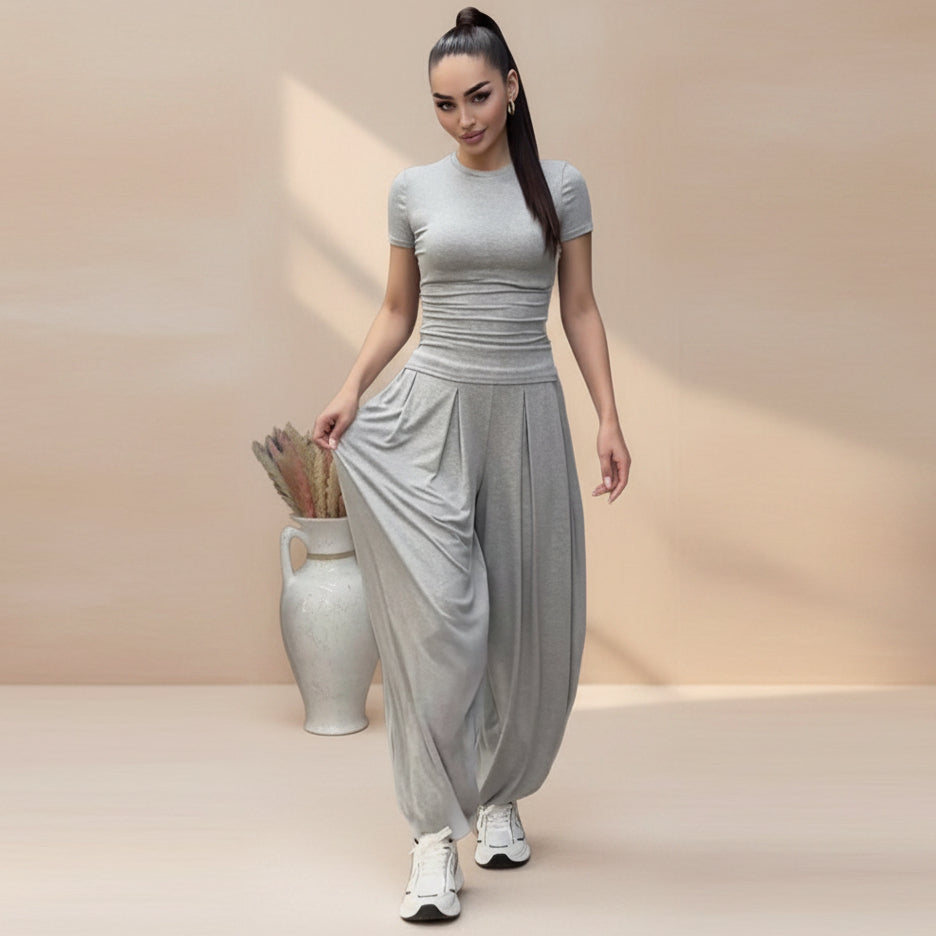 Model with a high ponytail holding out the wide leg of light gray pleated harem pants, paired with a matching short-sleeve fitted tee and white sneakers.