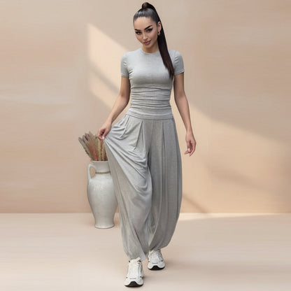 Model with a high ponytail holding out the wide leg of light gray pleated harem pants, paired with a matching short-sleeve fitted tee and white sneakers.